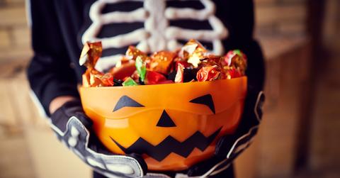 A person wearing a skeleton suit holding an orange bowl with a jack-o-lantern face full of candy.