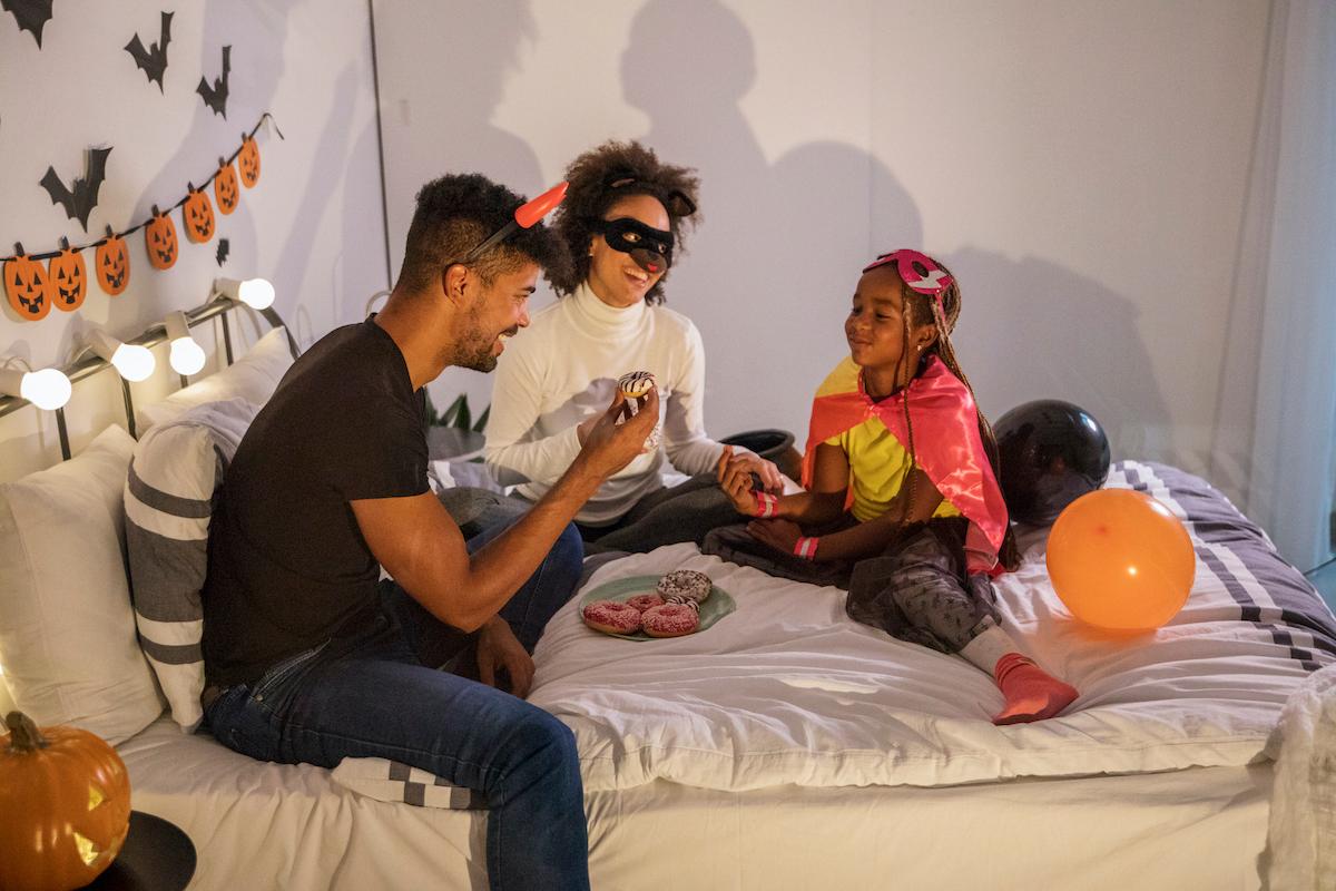 Parents and child eat donuts on a bed, wearing Halloween costumes