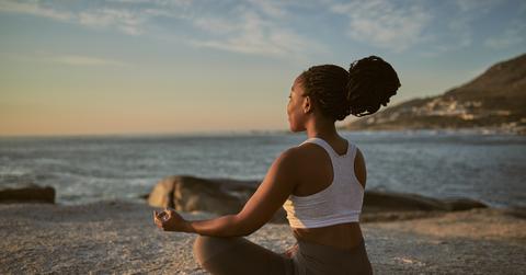 A young woman meditates while in a yoga pose atop a rock in a mountainous area overlooking the ocean.