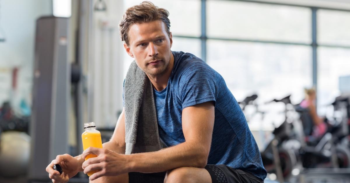 A man sits on a weight bench with a towel over his shoulder while drinking an orange sports drink.