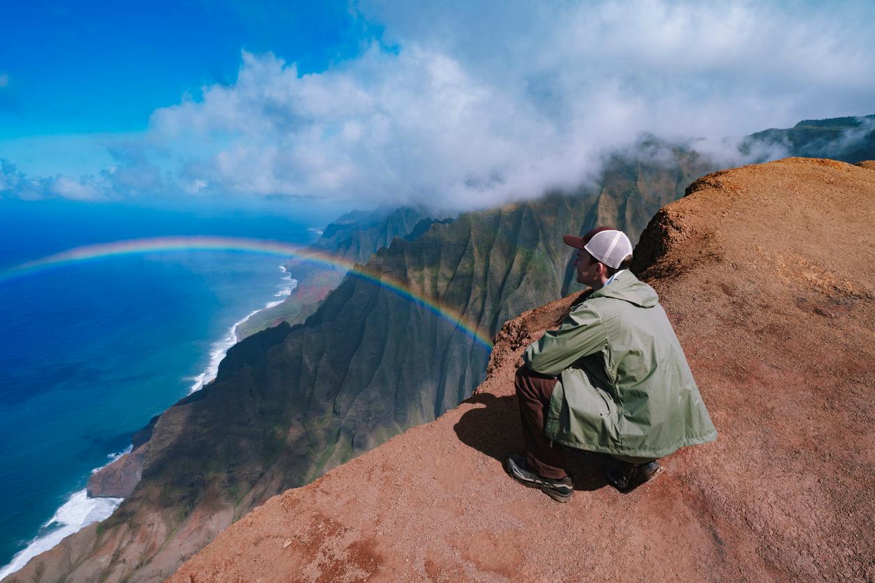 A hiker crouches above the edge of a cliff to view a rainbow beside a mountain with blue water below and cloudy skies above.