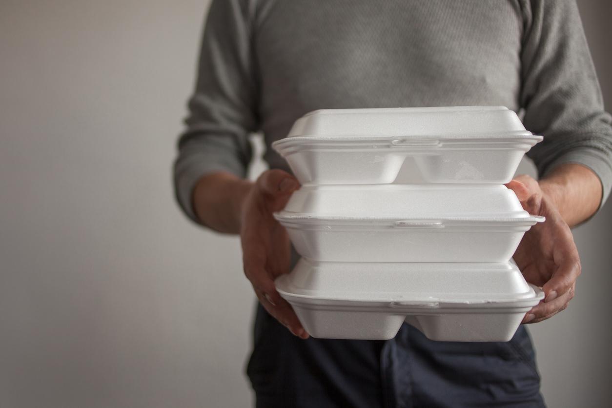 A man in a gray sweater holds three styrofoam takeout containers stacked atop one another.