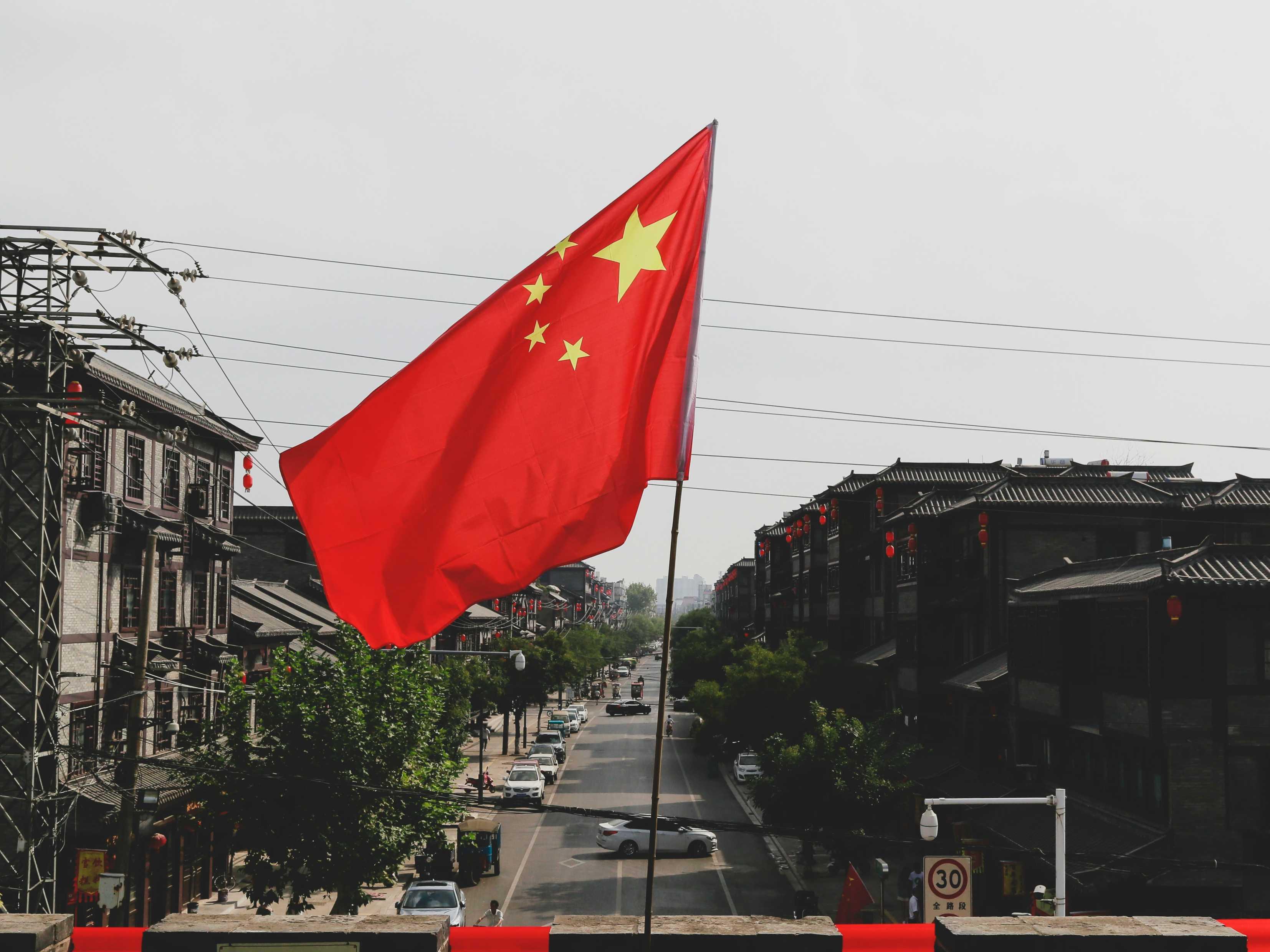 The flag of China is pictured in the center of a neighborhood with buildings on both sides and a tree-lined street in the center.