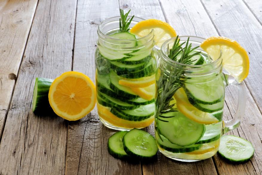 Two Mason jars of cucumber water garnished with rosemary and lemon atop a table next to cut cucumbers and lemons.