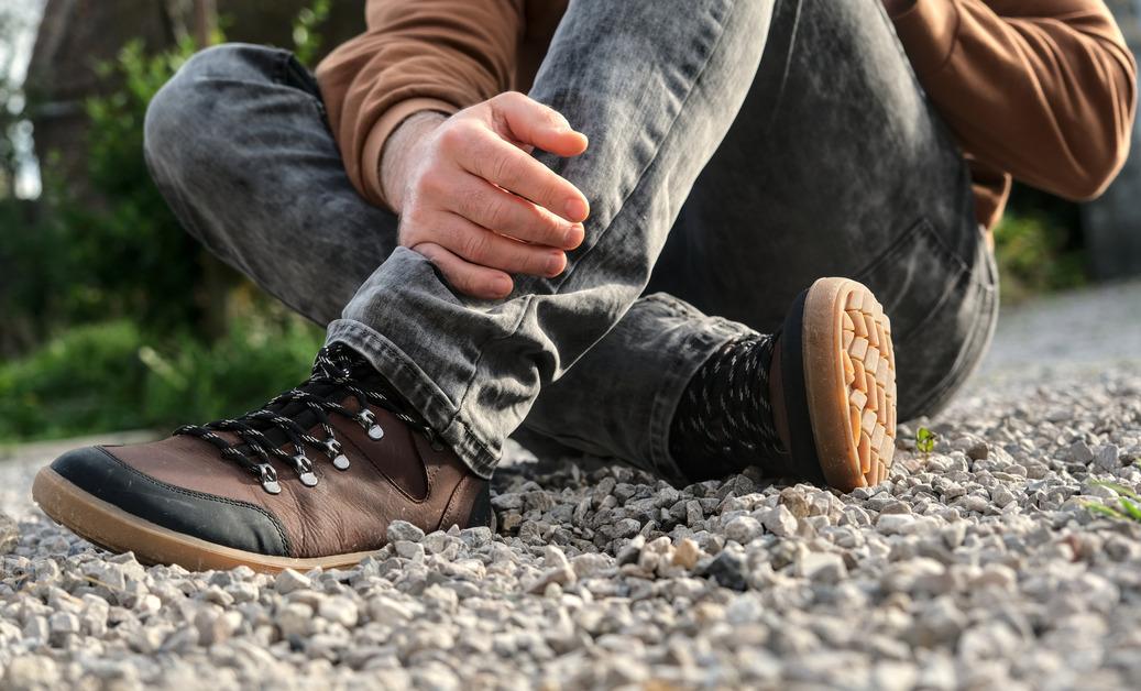 A person sits on gravel in gray jeans wearing a pair of minimalist brown shoes. 