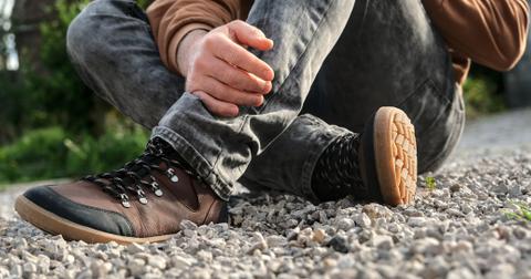 A person sits on gravel in gray jeans wearing a pair of minimalist brown shoes.
