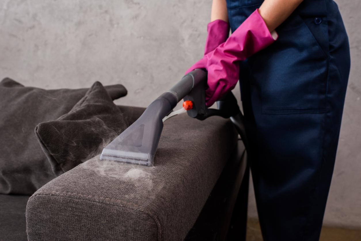 Close up of person using a vacuum attachment to vacuum the arm of a dark gray sofa.