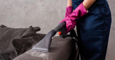 Close up of person using a vacuum attachment to vacuum the arm of a dark gray sofa.