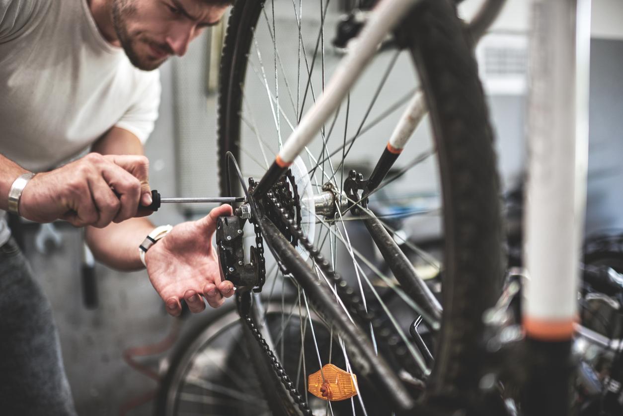 A man in a white shirt holds a screwdriver as he assembles a screw to hold a bike chain in place on a wheel.