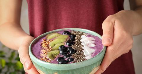 A woman in a red top holds an açaí smoothie bowl with fruit inside.
