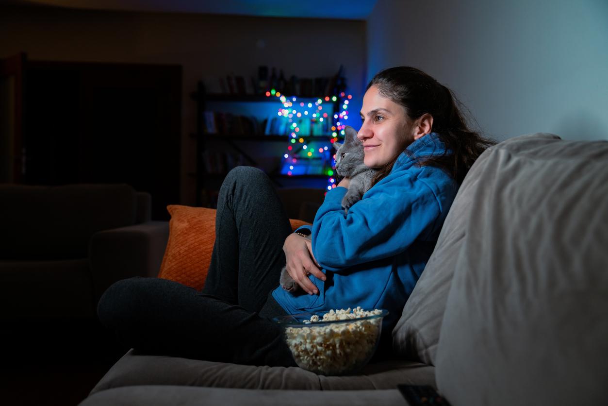 A woman in a blue hoodie cuddles a gray cat on a couch beside a glass bowl of popcorn.