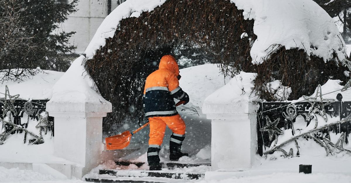 Person in a bright orange and black coat shovels his walkway beneath a snowy arch. 