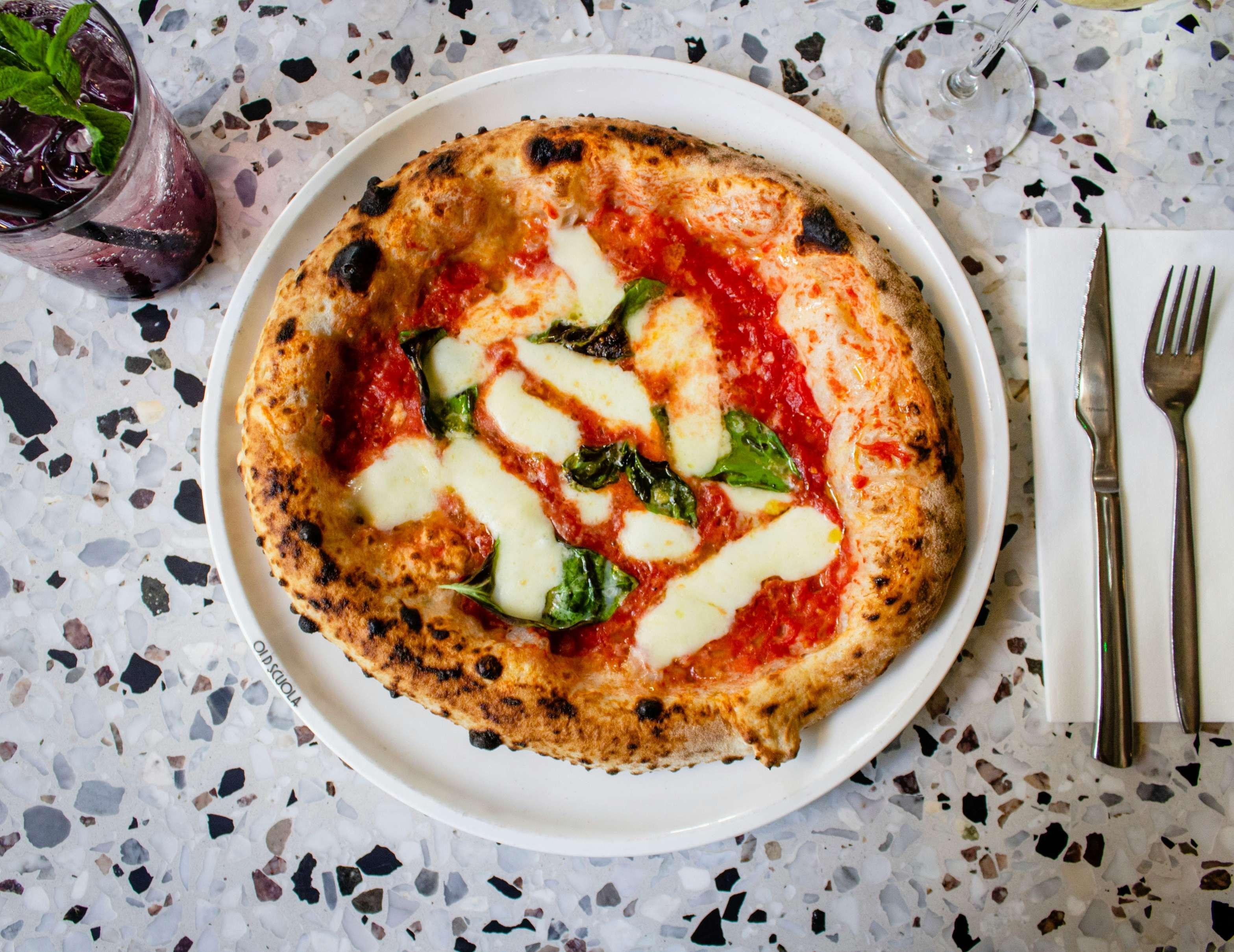 A close-up photo of a vegan margherita pizza on a plate with a drink in the corner and utensils on a napkin beside the plate.