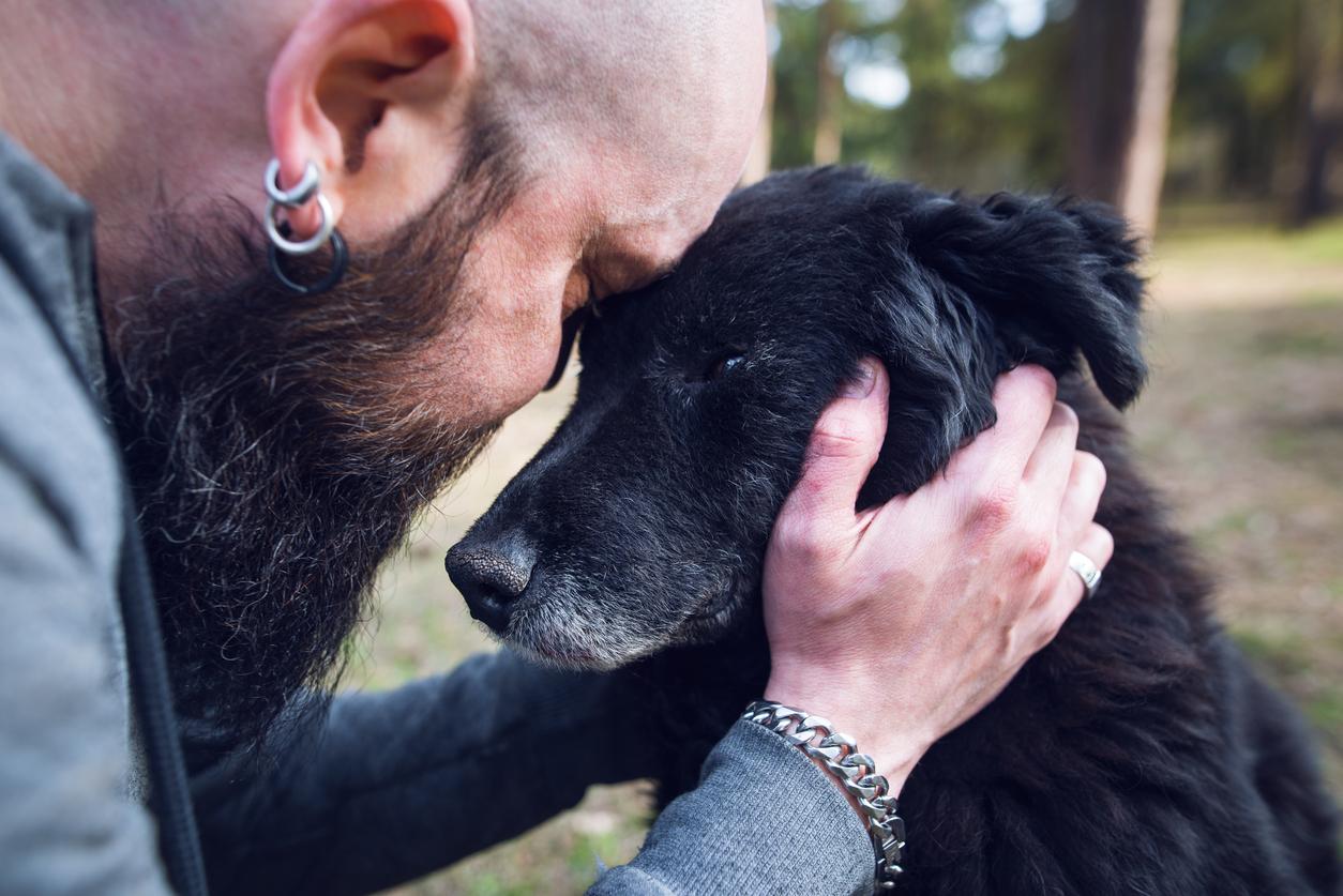 A man with earrings and a large beard comforts a black dog with their heads pressed against one another.