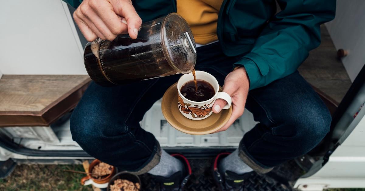 Person sitting on a bench pours coffee from a French press into a patterned mug on a white saucer