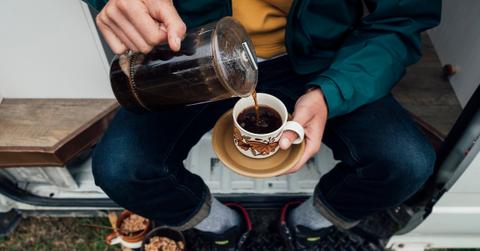 Person sitting on a bench pours coffee from a French press into a patterned mug on a white saucer