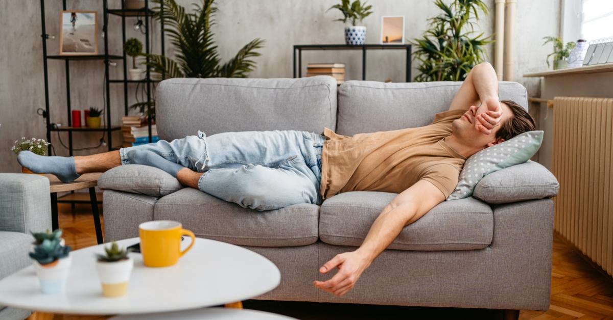 A man lies on a gray couch with his arm over his face. 