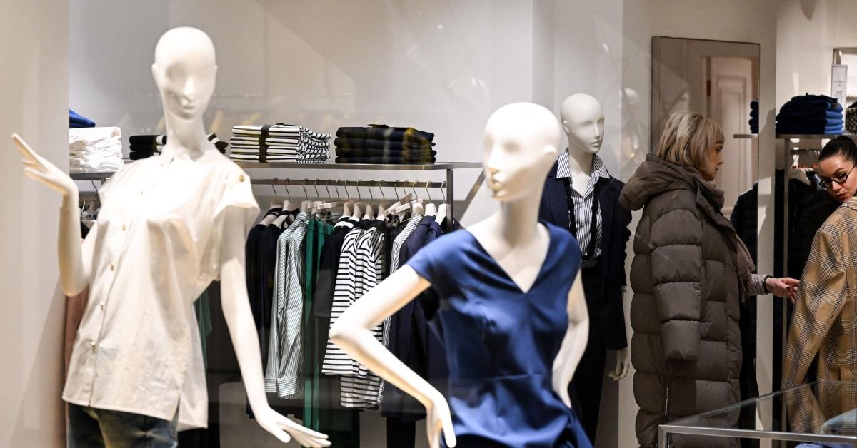 Two mannequins at the front of a clothing store, one wearing a blue shirt and the other a white shirt, with two women in the background shopping in the store. 