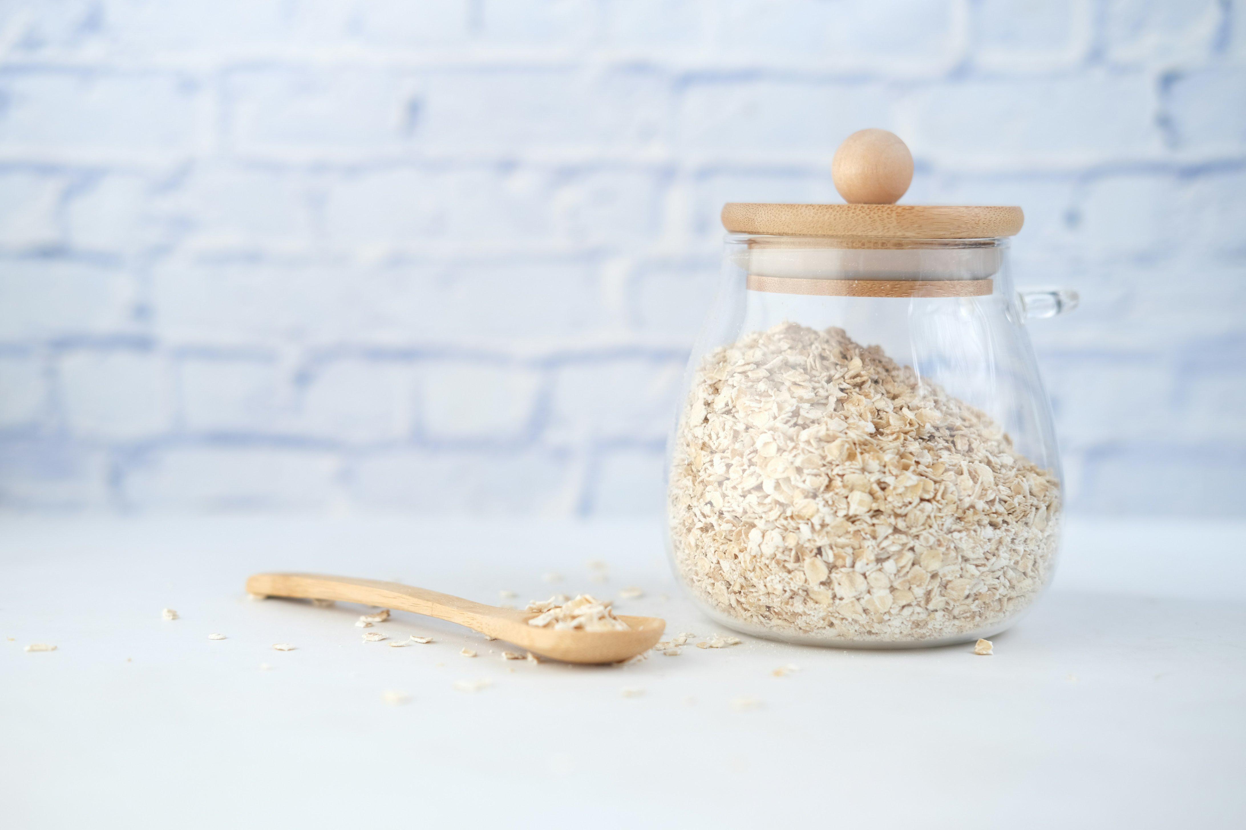 A glass jar of oats is pictured atop a counter beside a wooden spoon with oats in it.