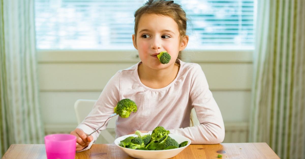 A pre-teen girl sitting at the dinner table eating broccoli. 