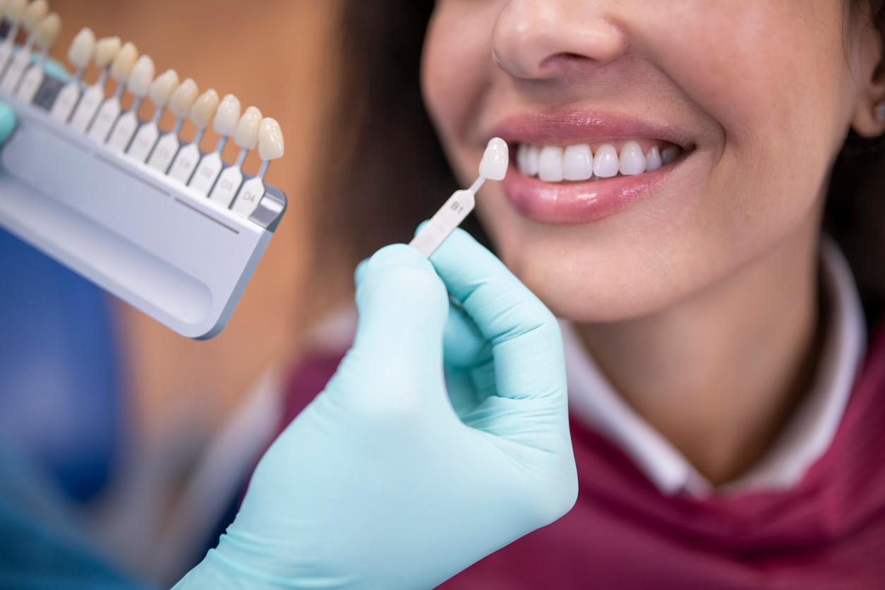 A dentist wearing blue gloves uses a palette tooth color sample to match the color of a patient's teeth.