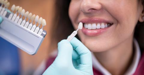 A dentist wearing blue gloves uses a palette tooth color sample to match the color of a patient's teeth.