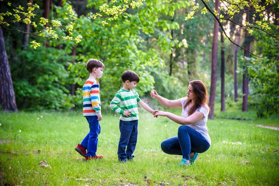 A mother sprays bug spray on her two sons while in the middle of the woods. 