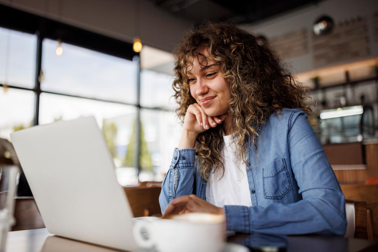 A young woman smiles down at her laptop while working inside a coffee shop.