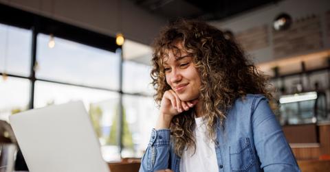 A young woman smiles down at her laptop while working inside a coffee shop.