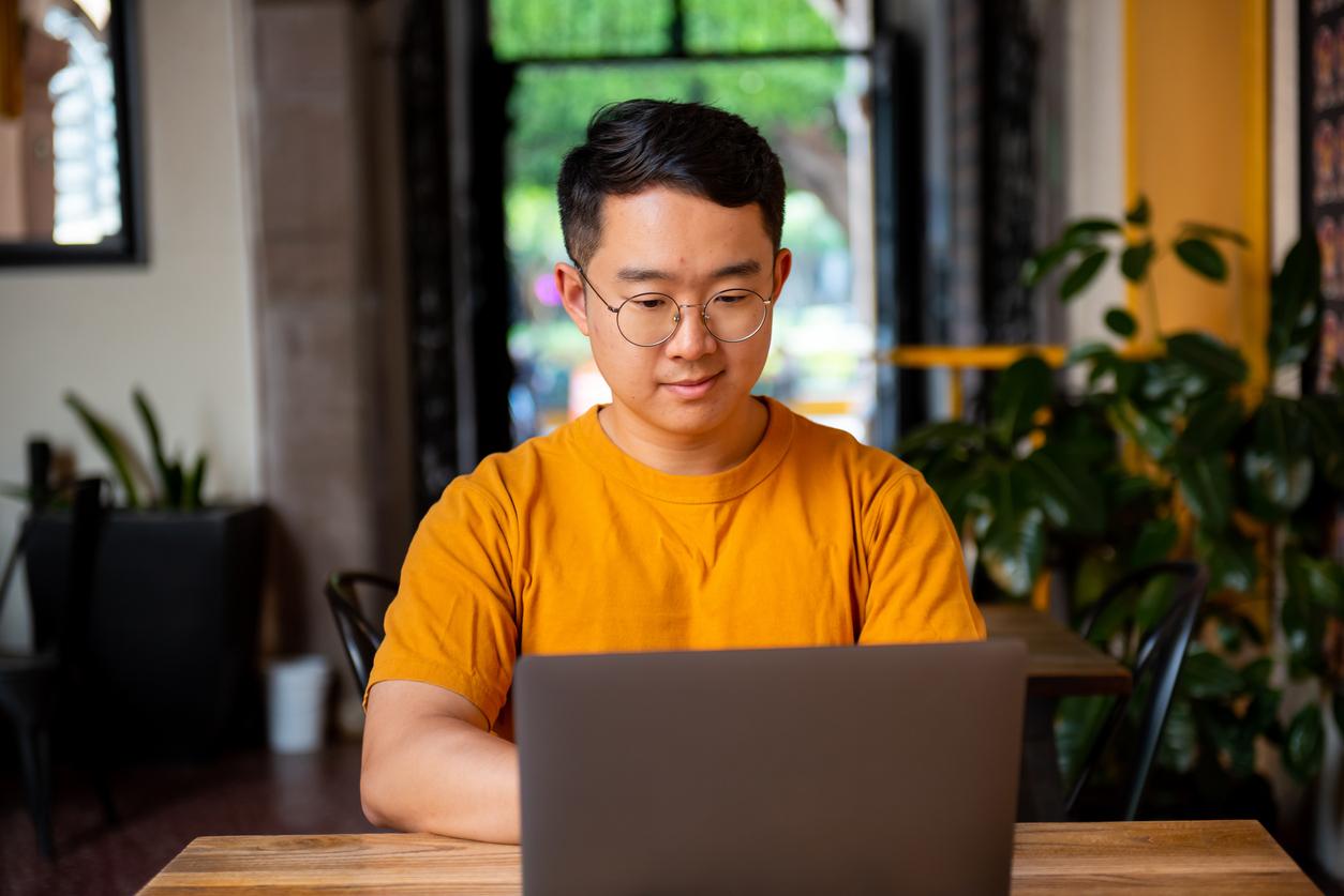 A young man in an orange shirt looks at his laptop while sitting in his living room with plants and curtains out of focus in the background.