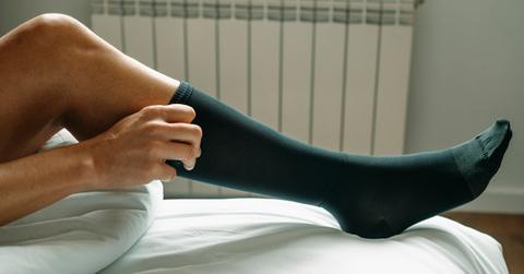 A man sitting on a bed putting on a dark green compression sock.