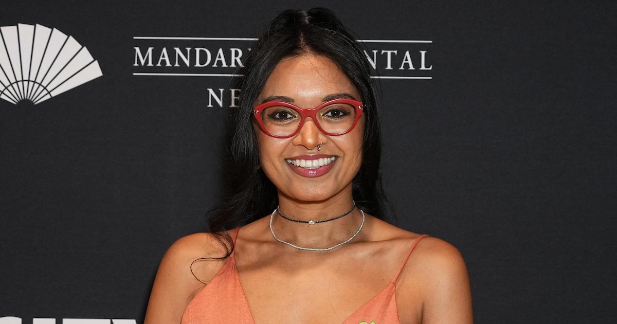 Priyanka Naik wears red glasses and an orange dress and smiles on a red carpet, at City Harvest Presents The 2024 Gala: Magic of Motown held at Cipriani 42nd Street on April 10, 2024 in New York City. 