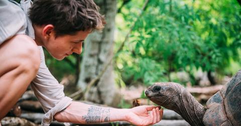 A young man in beige clothing extends his arm beneath a rescued turtle's head.