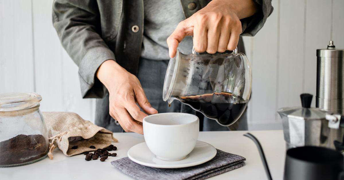 man pouring coffee into a cup and surrounded by grounds, beans, and coffee makers