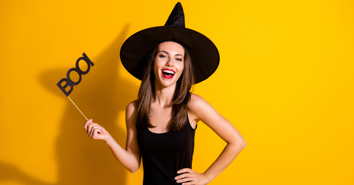 A woman holds a "boo" sign while dressed like a witch