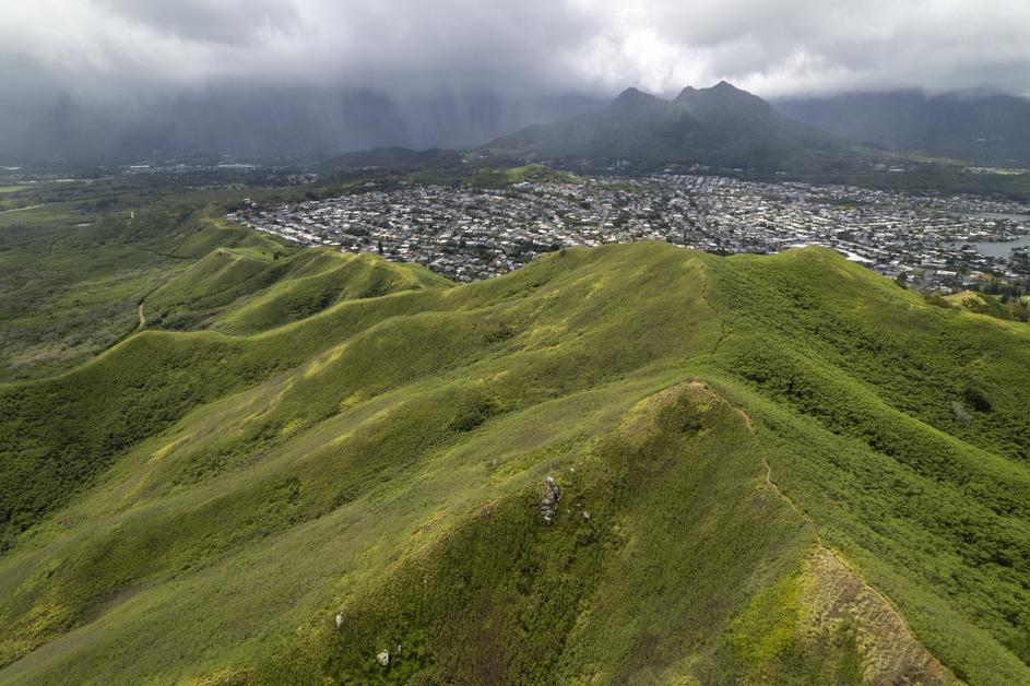 An aerial view of a lush green mountain in Oahu with the view of a city in the background.