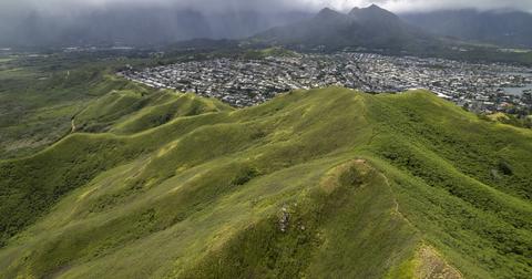 An aerial view of a lush green mountain in Oahu with the view of a city in the background.