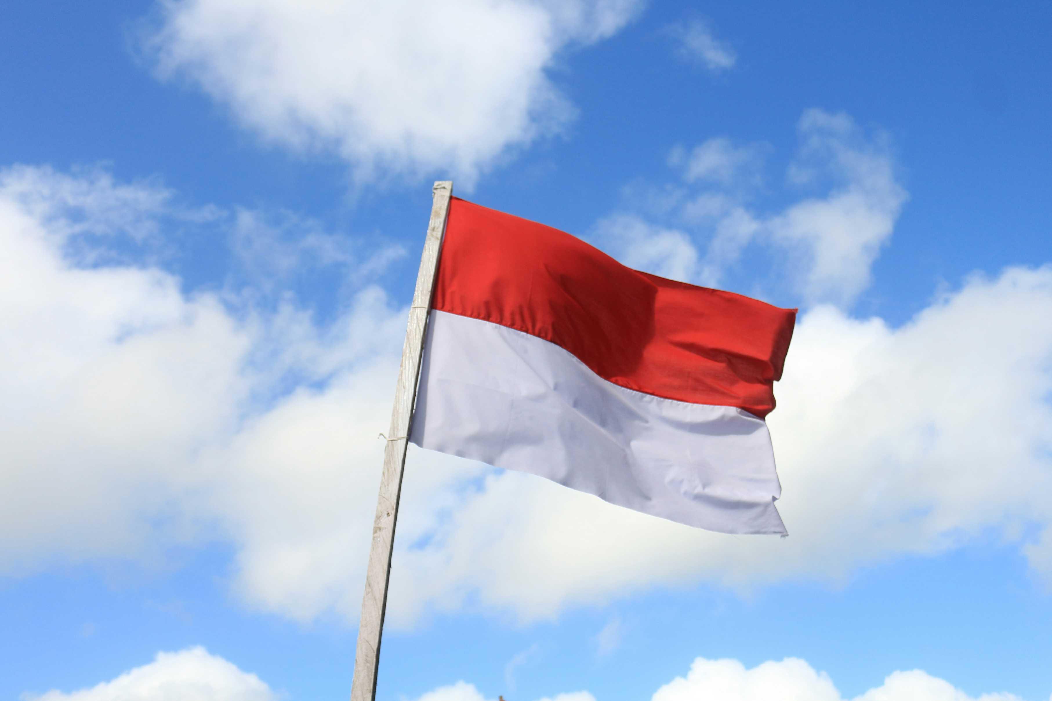 The flag of Indonesia is pictured waving on a pole against a cloudy blue sky.