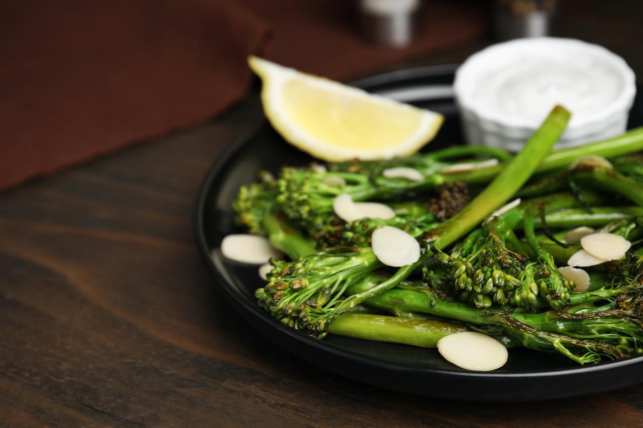 Close up of cooked broccoletti on a black plate with slivered almonds and a lemon wedge.