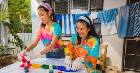 A mother and daughter tie dye shirts together on the patio beside their smiling dog.