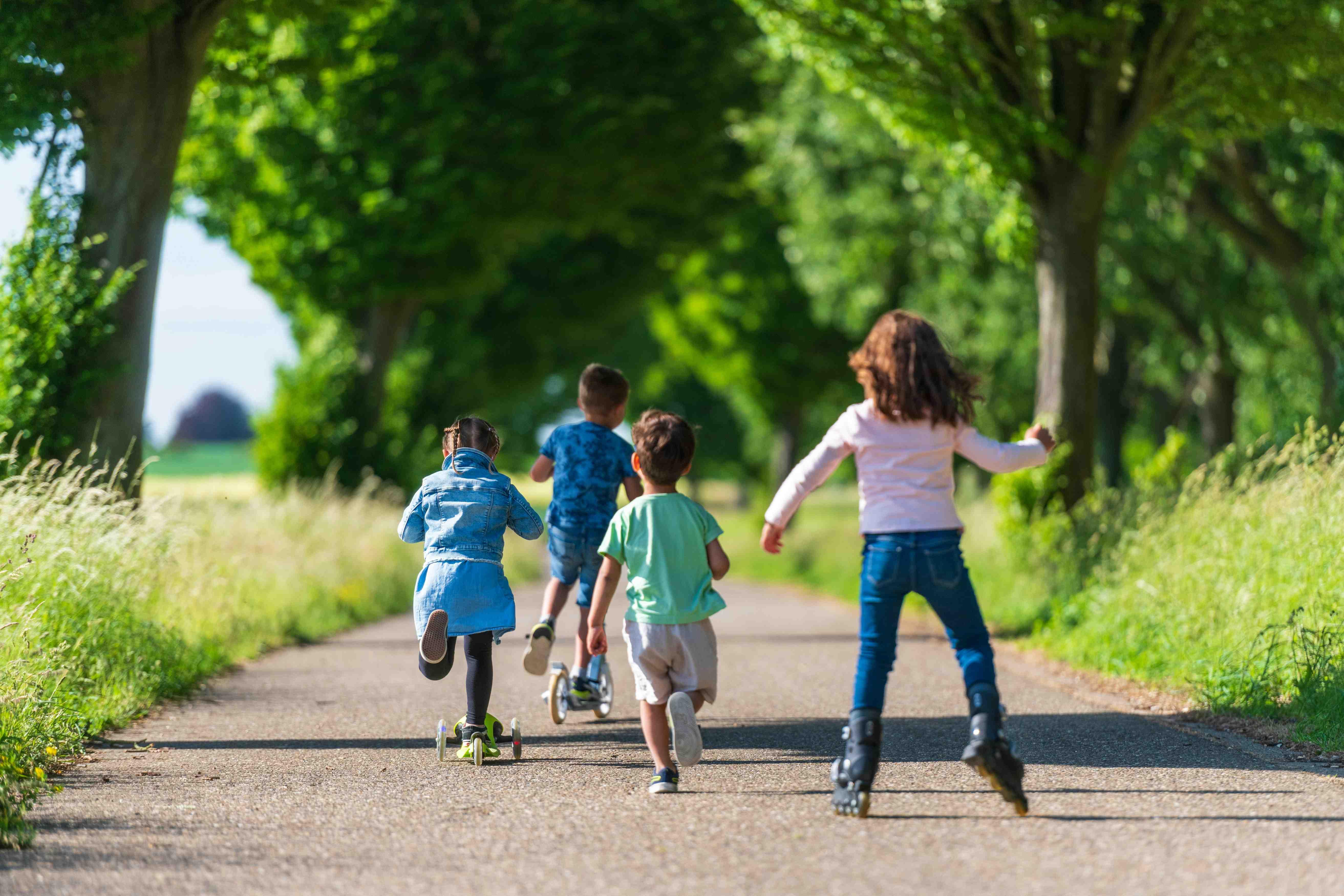 Several kids run and skate along a path surrounded by grass and trees.