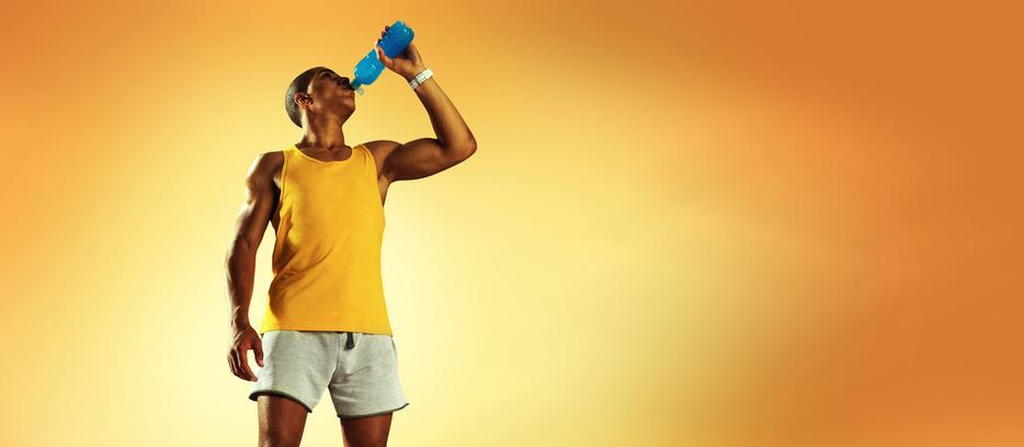 An athletic man wearing an orange tank top stands in front of an orange background while drinking from a blue bottle.