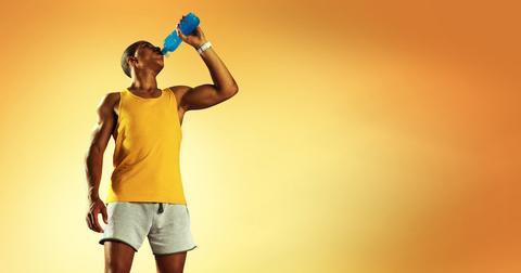 An athletic man wearing an orange tank top stands in front of an orange background while drinking from a blue bottle.