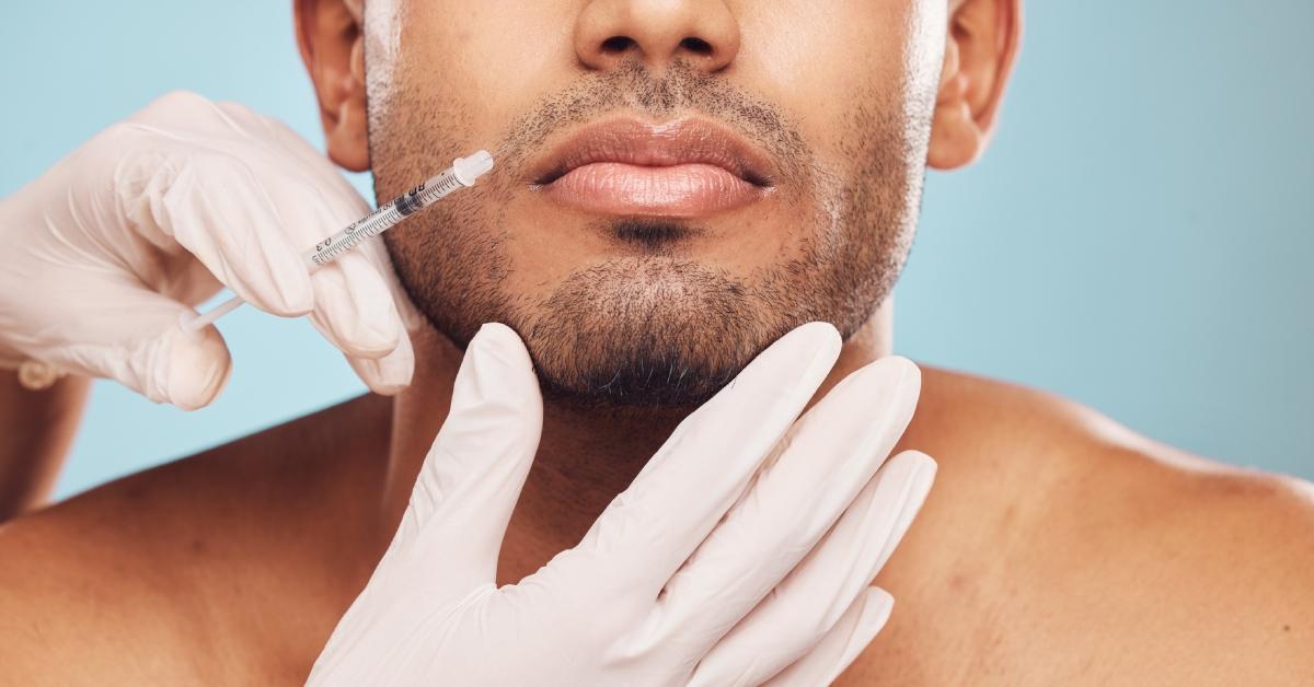 A pair of hands wearing white gloves holds a man's chin while putting an injection syringe up to his mouth. 