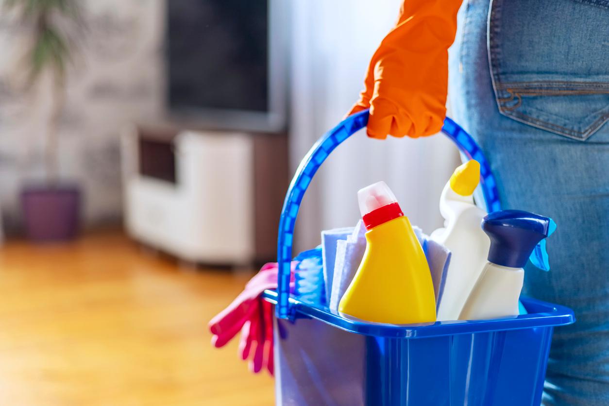 A cleaning professional holds a blue bucket with unlabeled containers of cleaning products in a living room.