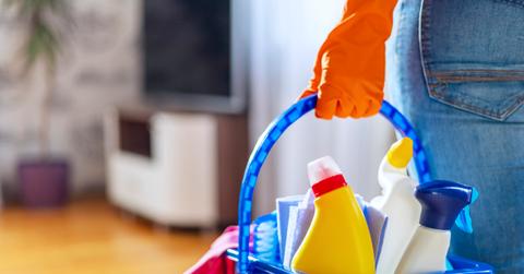 A cleaning professional holds a blue bucket with unlabeled containers of cleaning products in a living room.