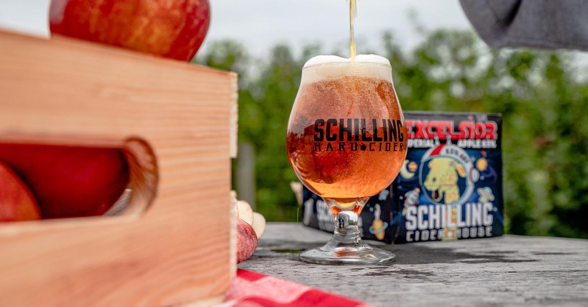 A glass of Schilling Cider sits next to a box of apples and a box of cider in an orchard.