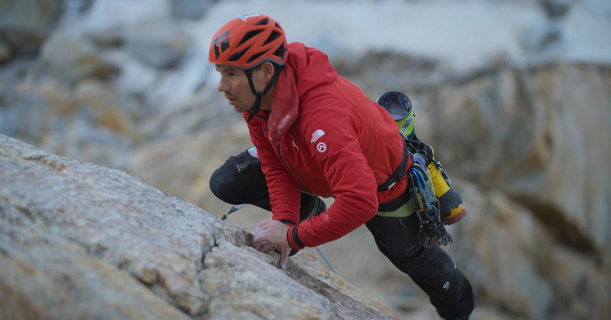 Alex Honnold in an orange helmet and red jacket, climbing a mountain.