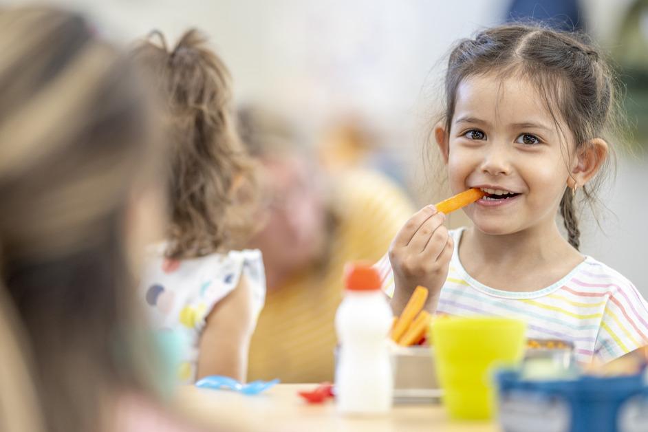 A little girl smiles while eating a carrot with the side of a girl's head in the foreground. 