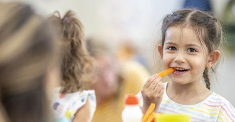 A little girl smiles while eating a carrot with the side of a girl's head in the foreground.